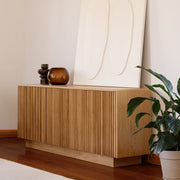 A light oak wood sideboard cabinet with vertical fluted detailing, styled in a minimalist room with a large cream abstract canvas leaning behind it, decorative objects on top, and a potted green plant on a dark wood floor.