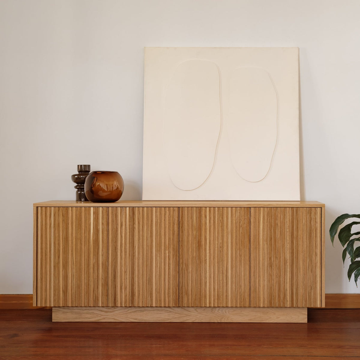 A mid-century modern wooden sideboard cabinet with vertical fluted wood-grain doors, styled with a large minimalist cream canvas print and small decorative vases, set against a neutral off-white wall and warm wood floor.