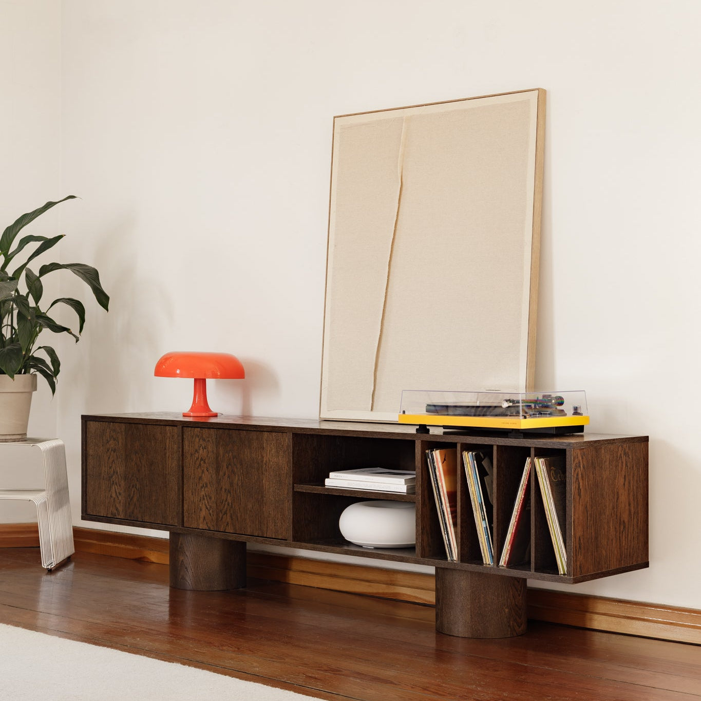 A mid-century modern wooden media console in a warm walnut finish, styled in a minimalist living room with a potted plant, orange table lamp, framed abstract art, and a yellow vinyl record player.