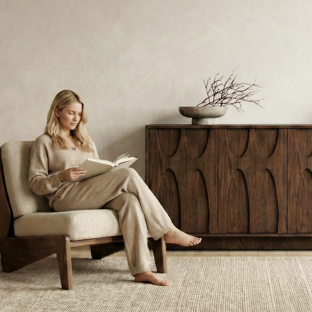 Woman reading a book in a cozy room with wooden furniture and a vase on a sideboard.